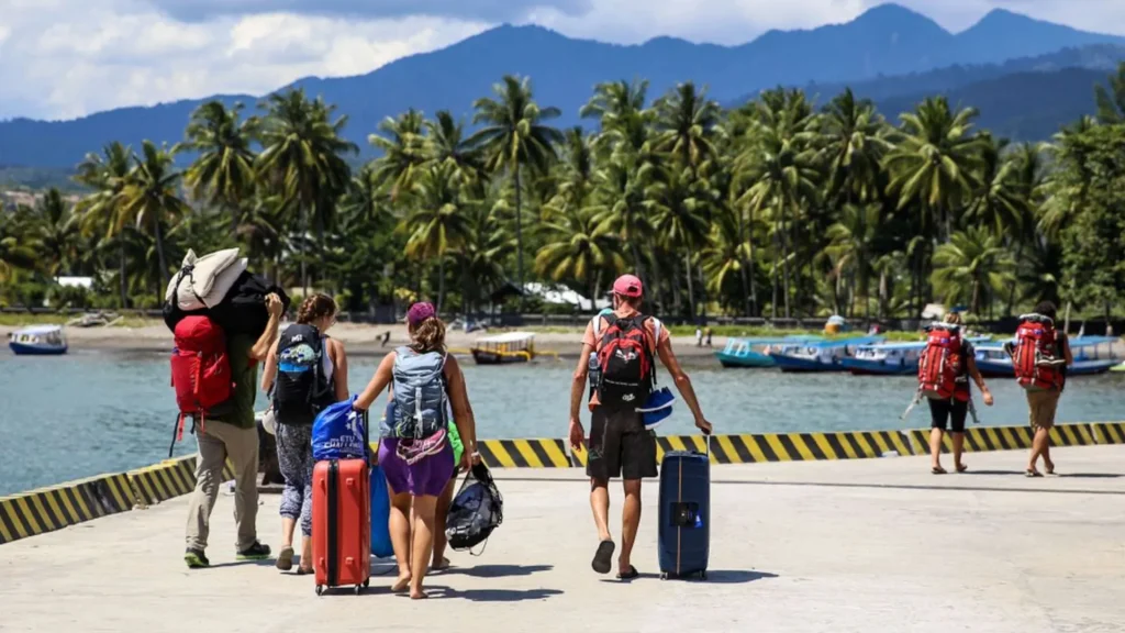 Backpackers carrying luggage and walking along the beach towards the embarkation area in the Bangsal port travel guide.